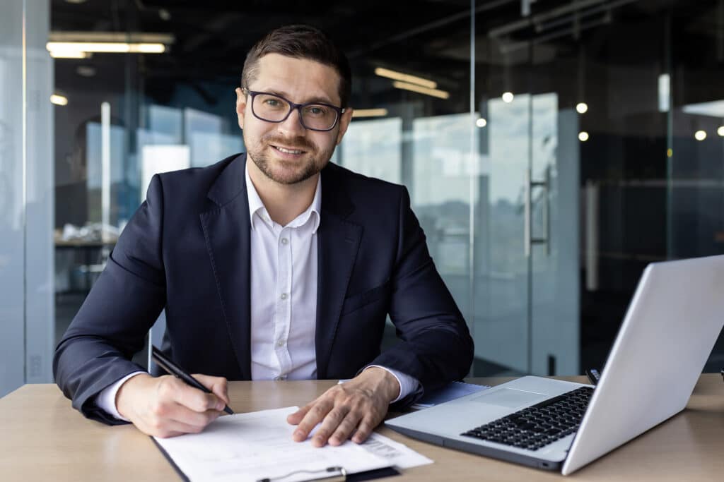 Portrait of successful financier accountant doing paperwork inside office, young man smiling and looking at camera in business suit